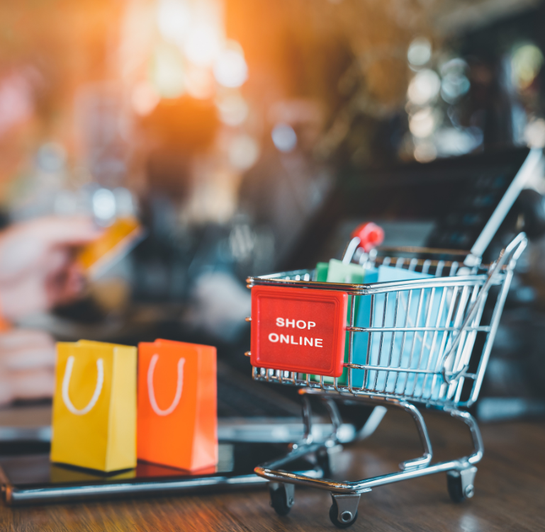 A shallow depth of field, wide-angle shot, featured in the file commerce-seo-services.jpg, of a miniature, metal shopping cart, filled with smaller gift bags in shades of blue and white, which features a red banner with the white, capitalized text "SHOP ONLINE" positioned on a wooden table, alongside other shopping bags in shades of yellow and orange in front of a blurred background containing a person holding a credit card over a laptop on a wooden desk.